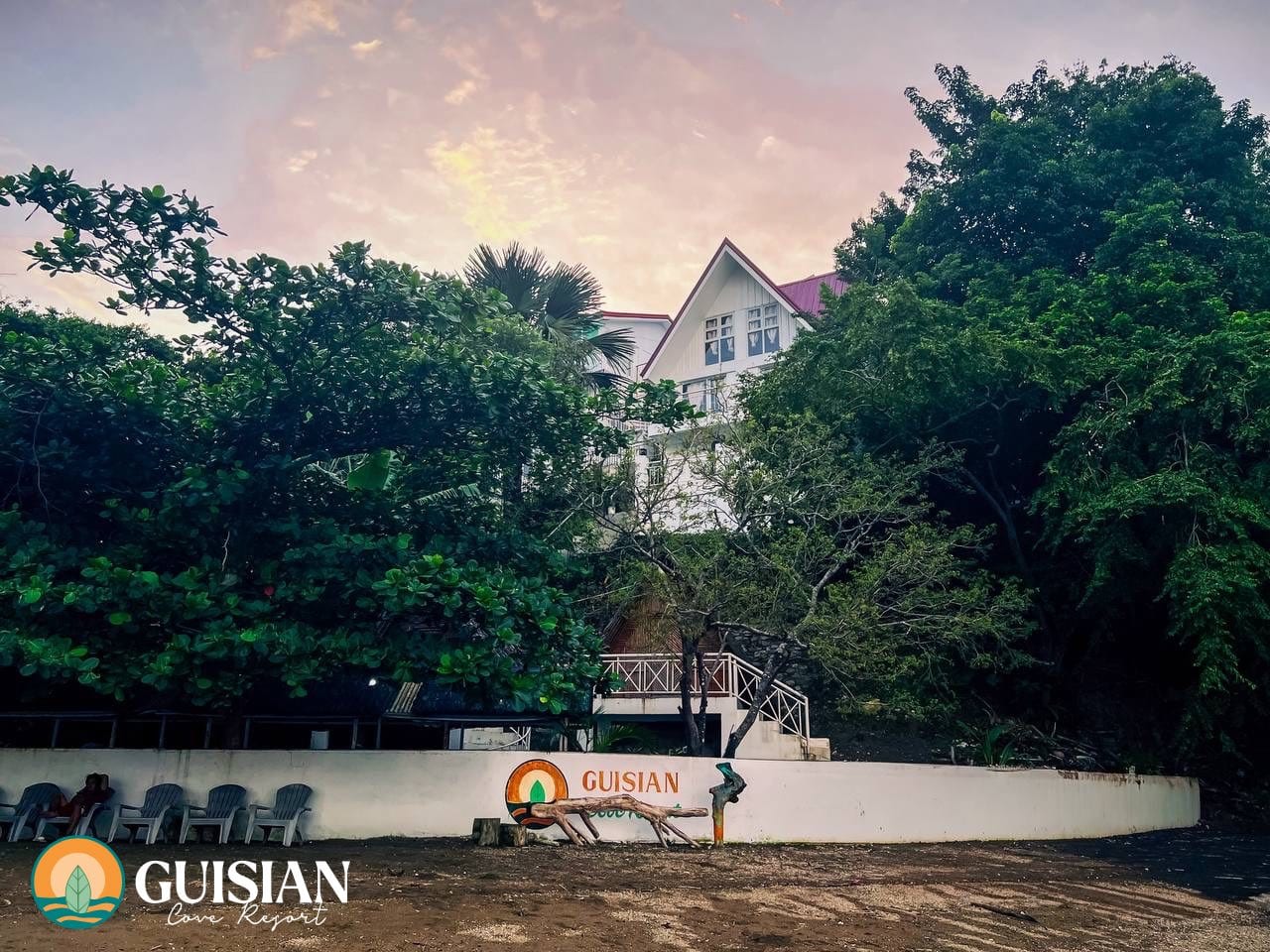 Beachfront view of Guisian Cove Resort at sunset, with the property's wooden signage on the sea wall and the main building rising above the trees